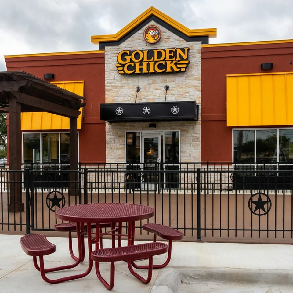 Golden Chick restaurant exterior with red and yellow facade, covered patio, black railings, and picnic table under cloudy skies.