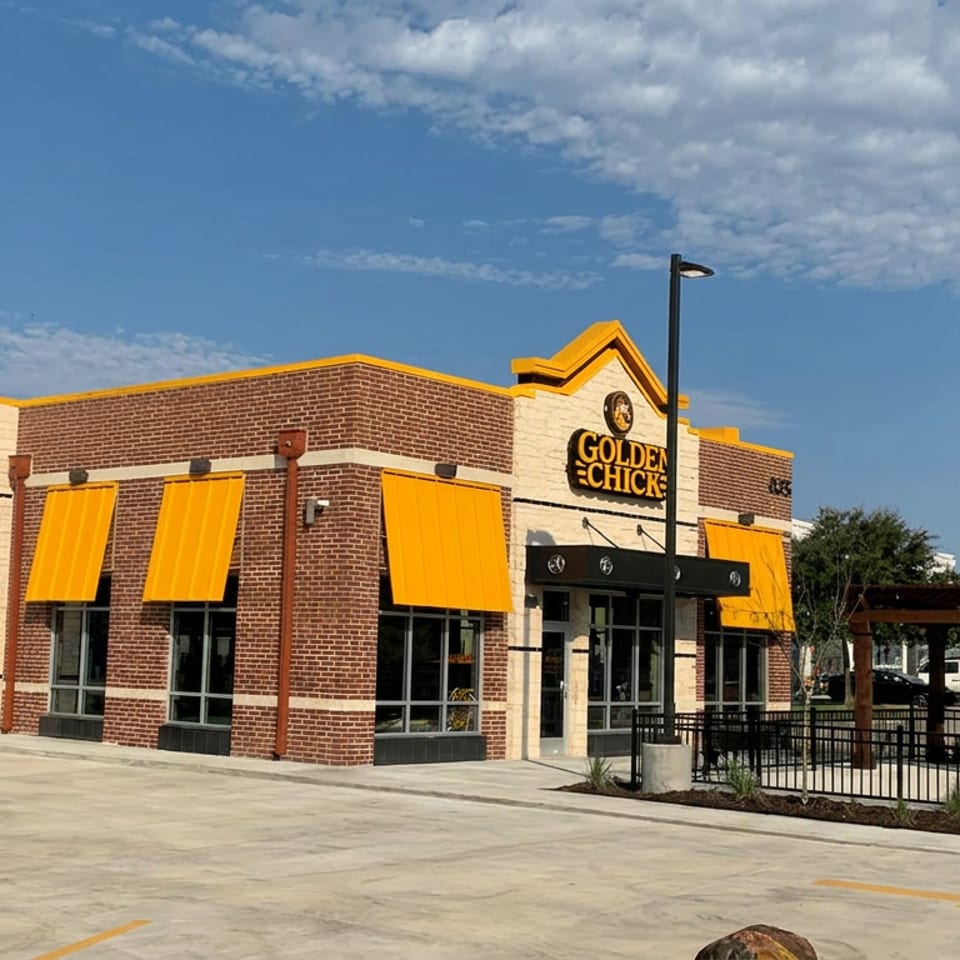 Golden Chick restaurant exterior with brick walls, yellow awnings, outdoor patio, and freestanding roadside sign under a blue sky.